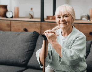 Seniorin sitzt lächelnd auf einem Sofa, genießt barrierefreies Wohnen und stützt sich auf einen Gehstock.